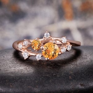 May include: A rose gold ring featuring a branch-like band set with two round orange citrine gemstones and several small, clear, round-cut white diamonds. The stones are arranged in a cluster along the top of the band, creating a delicate, organic appearance with a polished metal finish.