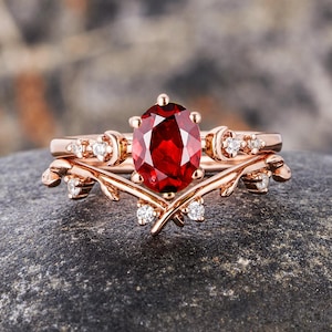 May include: Rose gold ring set featuring an oval-cut, deep red gemstone as the centerpiece. The engagement ring is accented with small, round, clear gemstones. The wedding band has a delicate, leaf-like design. The rings are displayed on a dark gray stone.