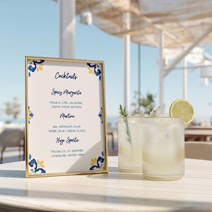 May include: A gold-framed cocktail menu with blue and yellow floral accents lists "Spicy Margarita," "Martini," and "Huge Spritz." Two clear, ribbed glasses with cocktails and ice sit on a table, garnished with lime and rosemary.