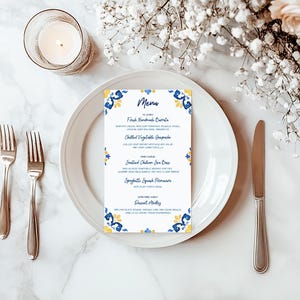 May include: A white plate with a menu card featuring blue and yellow floral designs. The menu lists various dishes. Silverware and a lit candle in a glass holder are on a marble surface, with white flowers in the background.