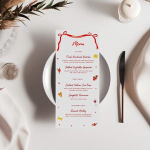 May include: A white menu card with red ribbon accents and the word "Menu" in elegant script. The menu lists dishes like burrata, gaspacho, sea bass, and spaghetti. A knife, plate, and napkin are also visible.