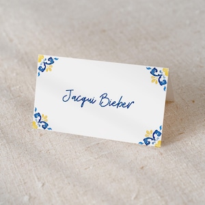 May include: A white place card with the name "Jacqui Bieber" in blue script. The corners are decorated with blue and yellow floral patterns. The card is displayed on a textured, neutral-colored surface.