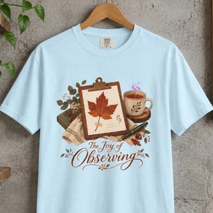 Könnte beinhalten: Ein hellblaues T-Shirt mit einer Grafik, die ein Klemmbrett mit einem Ahornblatt, eine Teetasse, einen Füllfederhalter und ein Buch zeigt. Der Text "The Joy of Observing" ist in brauner Schreibschrift unter dem Motiv aufgedruckt.