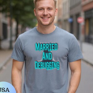 Könnte beinhalten: Ein graues T-Shirt mit dem Text "MARRIED AND DEBUGGING" in einem türkis-schwarzen Design. Die Person trägt eine schwarze Uhr. Das Bild ist im Freien aufgenommen.