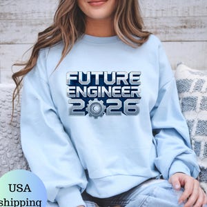May include: Pale blue sweatshirt featuring the text "FUTURE ENGINEER 2026" in a metallic, 3D-style font, with a gear graphic. The long-sleeved top is likely made of a soft fabric, suitable for casual wear.