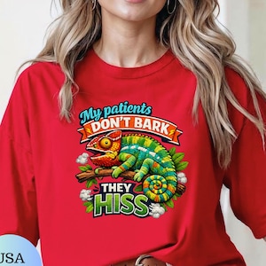 May include: A bright red t-shirt featuring a colorful graphic of a chameleon perched on a branch. The text reads My patients DON'T BARK THEY HISS in bold, stylized lettering. The design includes green leaves and white cloud-like accents.
