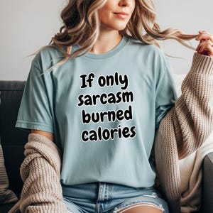 An individual sitting comfortably indoors wearing a casual short-sleeve graphic tee. The front displays the bold, playful text "If only sarcasm burned calories". This humorous workout shirt is an ideal funny gym t-shirt for fitness lovers who appreciate a good laugh during their exercise routines.