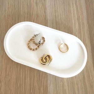 Może przedstawiać: Biała, owalna taca z trzema złotymi elementami biżuterii. Znajdują się na niej dwie pary kolczyków: kolczyki koła ze skręconym wzorem i małe, okrągłe kolczyki koła. Obecny jest również pojedynczy złoty nausznik.