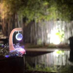 Puede incluir: Una bola de discoteca iluminada cuelga de un soporte blanco curvado. El soporte est&aacute; iluminado con luces LED de colores. El fondo presenta una escena exterior borrosa con &aacute;rboles y agua, creando un ambiente vibrante y festivo.