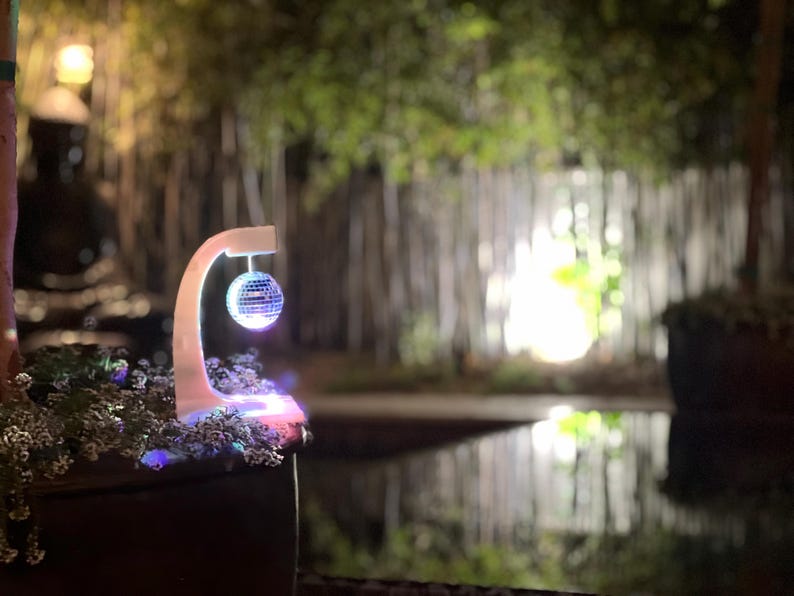 Puede incluir: Una bola de discoteca iluminada cuelga de un soporte blanco curvado. El soporte est&aacute; iluminado con luces LED de colores. El fondo presenta una escena exterior borrosa con &aacute;rboles y agua, creando un ambiente vibrante y festivo.