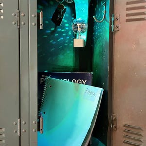 Puede incluir: Un casillero met&aacute;lico entreabierto, que revela un cuaderno turquesa con la etiqueta "English" y una peque&ntilde;a bola de discoteca. El interior del casillero est&aacute; iluminado con luz verde, y un llavero cuelga de un gancho.