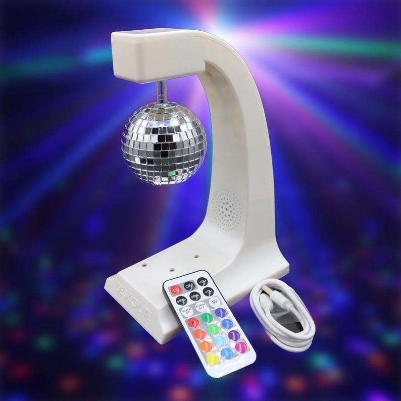 Puede incluir: Una luz de bola de discoteca blanca con una bola de espejo plateada suspendida de un soporte curvo. Se incluye un mando a distancia con botones de colores y un cable USB blanco. La base tiene el texto "TEK ROUTE".