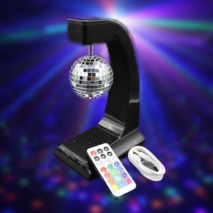 Puede incluir: Un soporte negro arqueado sostiene una bola de discoteca plateada. Un mando a distancia con botones coloridos y un cable USB blanco est&aacute;n delante del soporte. El fondo tiene un efecto de luz azul y p&uacute;rpura.