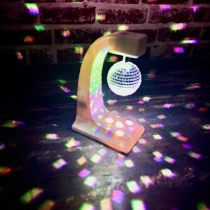 Puede incluir: Una peque&ntilde;a l&aacute;mpara de bola de discoteca beige con un brazo curvo y una base cuadrada. La l&aacute;mpara proyecta patrones de luz coloridos sobre la superficie circundante y una pared de ladrillos. La bola de discoteca es blanca con peque&ntilde;os cuadrados reflectantes.