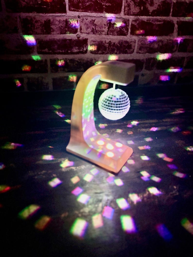 Puede incluir: Una peque&ntilde;a l&aacute;mpara de bola de discoteca beige con un brazo curvo y una base cuadrada. La l&aacute;mpara proyecta patrones de luz coloridos sobre la superficie circundante y una pared de ladrillos. La bola de discoteca es blanca con peque&ntilde;os cuadrados reflectantes.