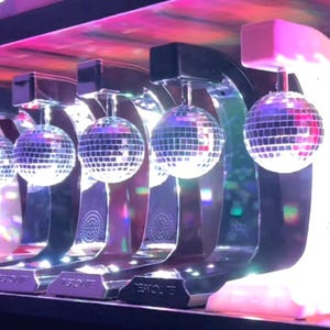 Op de afbeelding: Een rij discobol-lampen met een zilveren afwerking, hangend aan gebogen, verlichte standaards. De discobollen zijn rond en bedekt met kleine, reflecterende vierkantjes. De standaards hebben een rechthoekige basis met het woord "TESTOLITE" erop gedrukt.
