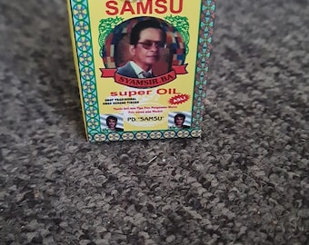 Può includere: Una piccola scatola gialla con la scritta "SAMSU" e "super OIL" in rosso e nero. La scatola presenta un ritratto di un uomo con gli occhiali e uno striscione con la scritta "SYAMSIR BA". Su una superficie strutturata.