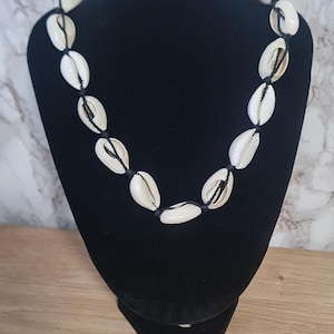 Peut inclure: Un collier composé d'une série de coquillages cauris blanc cassé enfilés sur un cordon noir fin. Les coquillages sont espacés uniformément le long de la longueur circulaire du cordon, créant un motif répétitif de coquillages marins naturels.