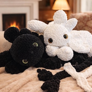 May include: Two plush dragon toys, one black and one white, rest on a soft, peach-colored blanket. The black dragon has green eyes, while the white dragon has brown eyes. The toys are handmade and have a soft, textured appearance.