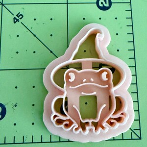 May include: A light peach-colored plastic cookie cutter shaped like a frog wearing a pointed hat. The cutter is positioned on a green cutting mat with black grid lines and the number 2 visible, showing the object is approximately 2 inches wide.