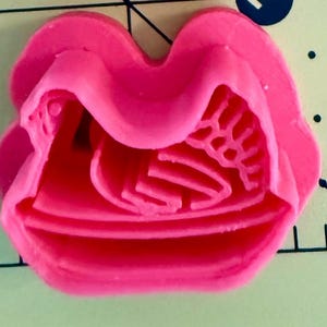 Könnte beinhalten: Ein hellrosa, rosenförmiger Ausstecher. Der Ausstecher ist aus Kunststoff gefertigt und weist detaillierte Merkmale auf, darunter Blütenblätter und ein Zentrum. Der Ausstecher befindet sich auf einer grünen Oberfläche mit schwarzen Linien.