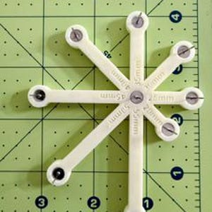 May include: A cream-colored, star-shaped tool with eight arms, each marked with millimeter measurements. The tool has small metal pins at the end of each arm and is set against a green grid background.