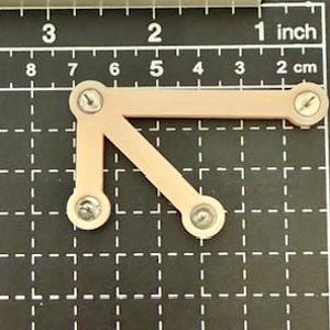 May include: A light tan, L-shaped, 3D-printed plastic bracket with three screws. The bracket is positioned on a black grid with inch and centimeter markings.