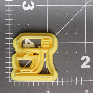 Può includere: Un tagliabiscotti giallo a forma di impastatrice planetaria posizionato su un tappetino da taglio con linee a griglia. L'oggetto misura circa 5 cm di larghezza e 4,5 cm di altezza, come indicato dai segni sulla griglia di misurazione.