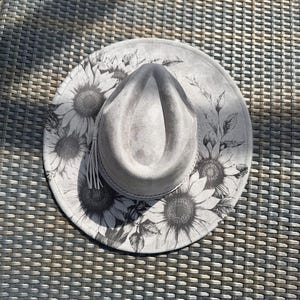 May include: A light grey fedora hat featuring a wide brim decorated with a black and white sunflower print. The hat has a pinched crown and a thin, braided cord band encircling the base of the crown, with a small tassel detail on the side.