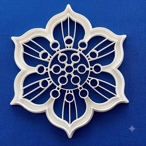 May include: A white plastic cookie cutter shaped like a six-petaled flower. The design features a central circular hub connected to six smaller circles, with each petal containing three radiating lines that extend toward the outer edge.