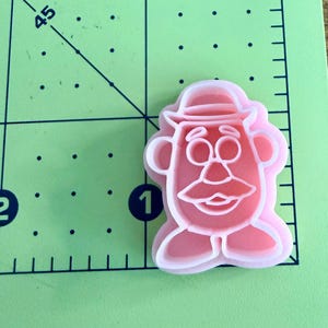 May include: A light pink plastic cookie cutter shaped like a potato character with a hat, mustache, and shoes. The cutter is positioned on a green cutting mat with a visible ruler showing a measurement of approximately 1.5 inches in width.
