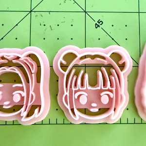 May include: Three light pink plastic cookie cutters shaped like stylized cartoon faces with different hairstyles, including pigtails, space buns, and a high ponytail, arranged on a green cutting mat with visible grid lines and numbers.