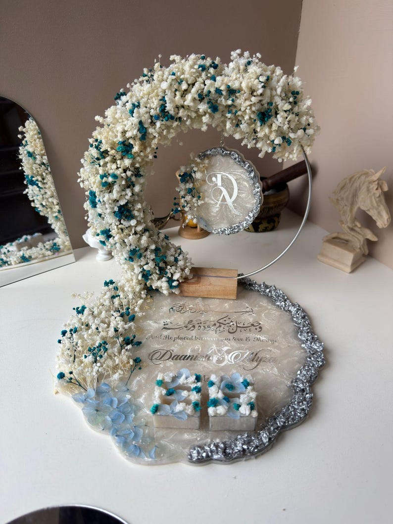 K&ouml;nnte beinhalten: Dekorative Hochzeits- oder Event-Tischdekoration mit einem Blumenbogen aus wei&szlig;en und t&uuml;rkisfarbenen Bl&uuml;ten. Die Basis ist ein Harztablett mit Text und zwei kleinen Boxen. Ein silberfarbener Metallrahmen tr&auml;gt den Bogen und ein kreisf&ouml;rmiges Emblem.