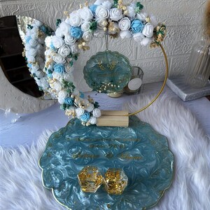 K&ouml;nnte beinhalten: Dekorative Hochzeitsausstellung mit einer tealfarbenen Harzschale mit goldenen Akzenten, die zwei goldgef&uuml;llte geometrische Boxen h&auml;lt. Ein goldener Reifen tr&auml;gt ein Blumenarrangement aus wei&szlig;en und blauen Rosen. Ein tealfarbenes Ornament h&auml;ngt.