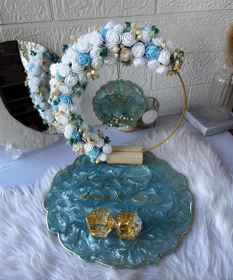 K&ouml;nnte beinhalten: Dekorative Hochzeitsausstellung mit einer tealfarbenen Harzschale mit goldenen Akzenten, die zwei goldgef&uuml;llte geometrische Boxen h&auml;lt. Ein goldener Reifen tr&auml;gt ein Blumenarrangement aus wei&szlig;en und blauen Rosen. Ein tealfarbenes Ornament h&auml;ngt.