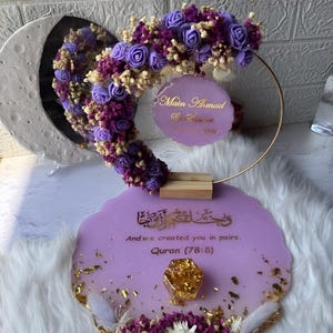 K&ouml;nnte beinhalten: Dekorative Tischdekoration f&uuml;r Hochzeiten oder Veranstaltungen mit lavendelfarbener Harzbasis, Goldblattakzenten und Blumenarrangements. Es umfasst einen goldenen Ring, einen Spiegel und ein Schild mit Text. Dekoriert mit lila und wei&szlig;en Blumen.