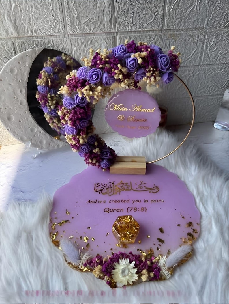 K&ouml;nnte beinhalten: Dekorative Tischdekoration f&uuml;r Hochzeiten oder Veranstaltungen mit lavendelfarbener Harzbasis, Goldblattakzenten und Blumenarrangements. Es umfasst einen goldenen Ring, einen Spiegel und ein Schild mit Text. Dekoriert mit lila und wei&szlig;en Blumen.