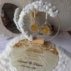 K&ouml;nnte beinhalten: Dekorative Hochzeitsdekoration mit einem goldfarbenen, muschelf&ouml;rmigen Tablett und einem Blumengesteck. Es umfasst zwei Glaskugeln mit Goldflocken, einen kreisf&ouml;rmigen Metallrahmen und wei&szlig;e Kunstblumen.