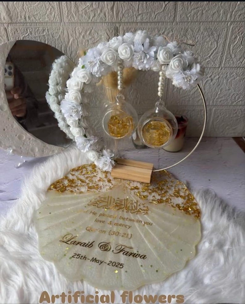 K&ouml;nnte beinhalten: Dekorative Hochzeitsdekoration mit einem goldfarbenen, muschelf&ouml;rmigen Tablett und einem Blumengesteck. Es umfasst zwei Glaskugeln mit Goldflocken, einen kreisf&ouml;rmigen Metallrahmen und wei&szlig;e Kunstblumen.