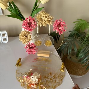 K&ouml;nnte beinhalten: Ein dekoratives Tablett mit goldenen Akzenten, das ein Blumenarrangement und zwei Glaskugeln zeigt. Das Tablett hat ein wei&szlig;es und goldenes Design mit goldenen Griffen. Das Arrangement umfasst rosa und beige Trockenblumen.