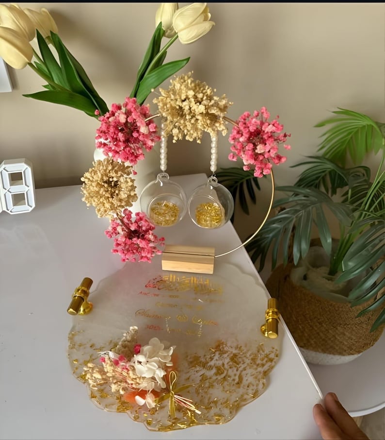 K&ouml;nnte beinhalten: Ein dekoratives Tablett mit goldenen Akzenten, das ein Blumenarrangement und zwei Glaskugeln zeigt. Das Tablett hat ein wei&szlig;es und goldenes Design mit goldenen Griffen. Das Arrangement umfasst rosa und beige Trockenblumen.