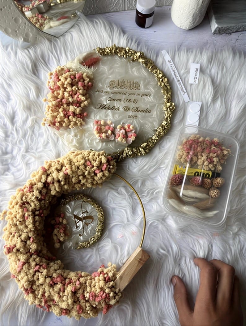 K&ouml;nnte beinhalten: Dekorative Hochzeits- oder Event-Dekoration mit klarer Acrylbasis, goldenen Verzierungen und floralen Akzenten in Rosa und Beige. Das Design umfasst ein rundes Element mit Text und eine spiralf&ouml;rmige Form. Der Text lautet "And we created you in pairs Quran (78.8) Abdullah @ Saadia 2020."