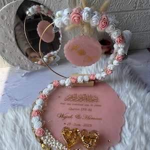 K&ouml;nnte beinhalten: Rosa-goldene Hochzeitsdekoration mit Spiegel, Blumenschmuck und dekorativer Plakette. Die Plakette enth&auml;lt arabischen und englischen Text sowie das Datum "21-June-2025". Zwei goldene herzf&ouml;rmige Verzierungen sind ebenfalls vorhanden.