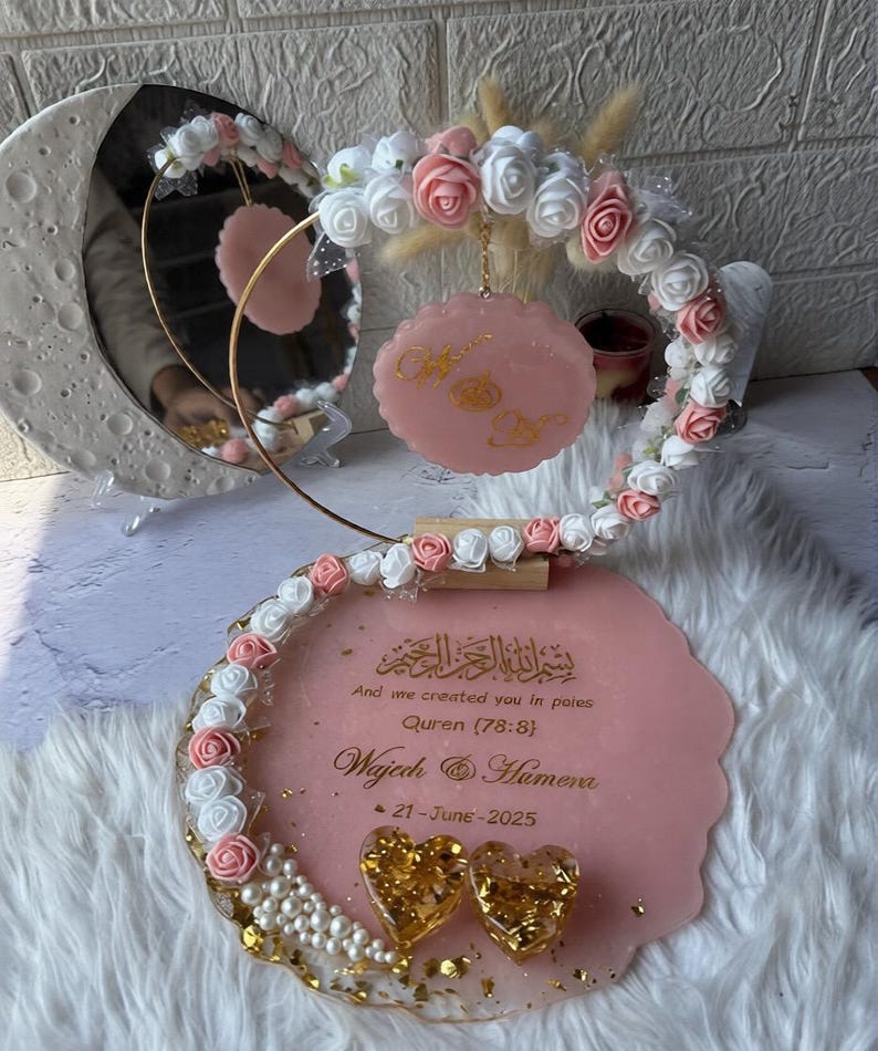 K&ouml;nnte beinhalten: Rosa-goldene Hochzeitsdekoration mit Spiegel, Blumenschmuck und dekorativer Plakette. Die Plakette enth&auml;lt arabischen und englischen Text sowie das Datum "21-June-2025". Zwei goldene herzf&ouml;rmige Verzierungen sind ebenfalls vorhanden.