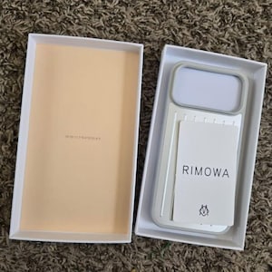 May include: A light gray Rimowa phone case with a rectangular camera cutout and a white insert card featuring the brand name Rimowa and a small logo, presented inside a white rectangular box with a light beige interior lining.
