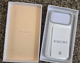 Può includere: Una custodia per telefono Rimowa grigio chiaro con un ritaglio rettangolare per la fotocamera e una scheda bianca con il nome del marchio Rimowa e un piccolo logo, presentata in una scatola rettangolare bianca con interno beige.