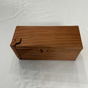 May include: A rectangular wooden box with a natural light brown grain pattern. The box features a sliding lid mechanism with a visible interlocking joint on the left side and a small carved indentation on the front edge for easy opening.