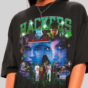 Op de afbeelding: Zwart oversized T-shirt met een grafisch ontwerp met het woord "Hackers" in groen en een collage van afbeeldingen. Het ontwerp bevat gezichten, sterren en tekst. Het shirt heeft korte mouwen.