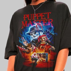 May include: Black t-shirt featuring a colorful graphic from the movie "Puppet Master." The design includes a skull in a hat, a jester, and other characters. The title "Puppet Master" is displayed in red and blue lettering.