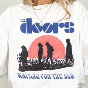 Op de afbeelding: Wit XL T-shirt met een afbeelding van de band The Doors. Het ontwerp bevat de bandnaam in blauw, een rode zon en silhouetten van mensen. De tekst "WAITING FOR THE SUN" staat eronder.